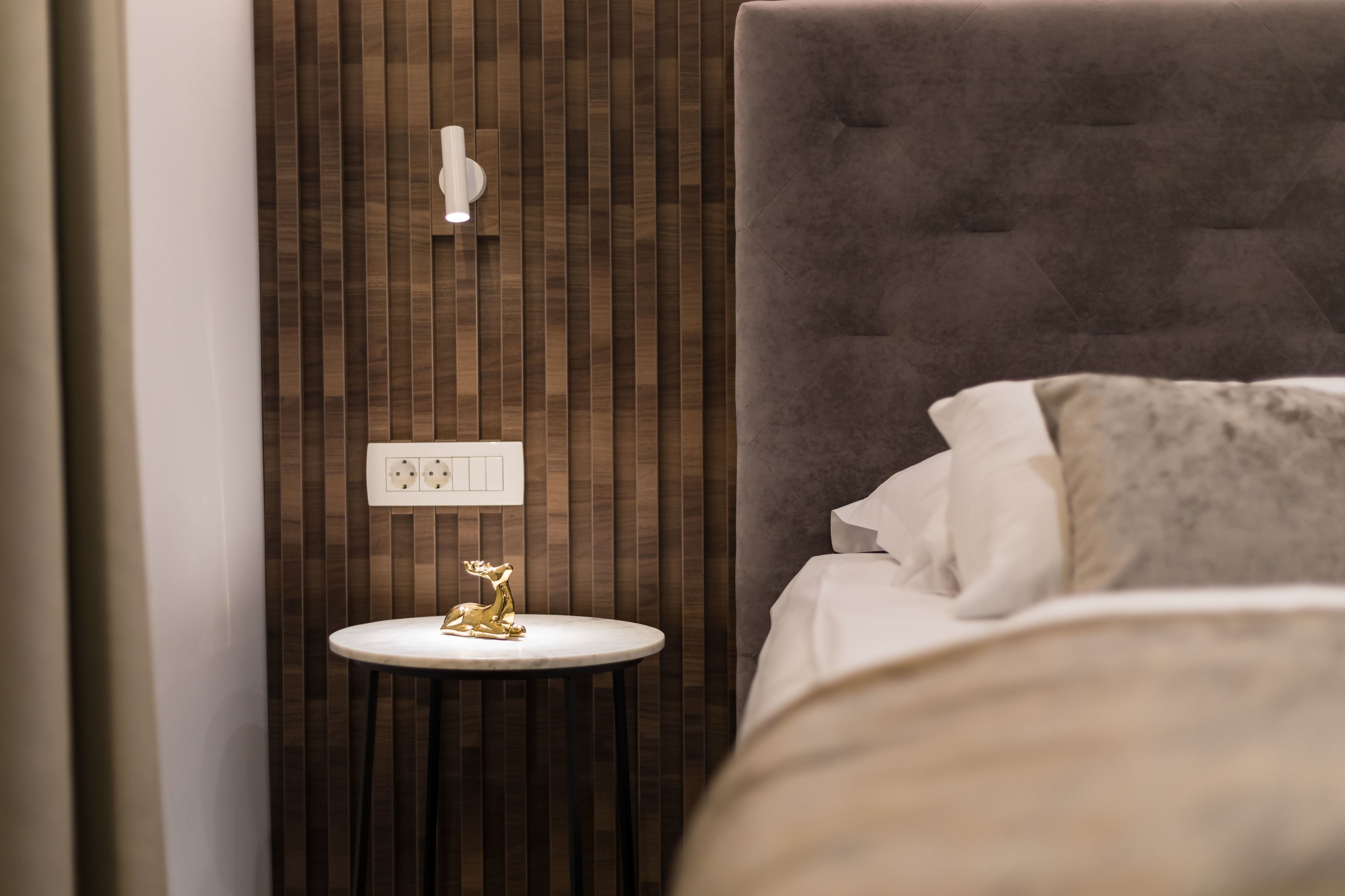 Ein Schlafzimmer mit einem Bett, einem runden weißen Nachttisch mit einer goldenen Statue, einer Steckdose und einer Wandleuchte.