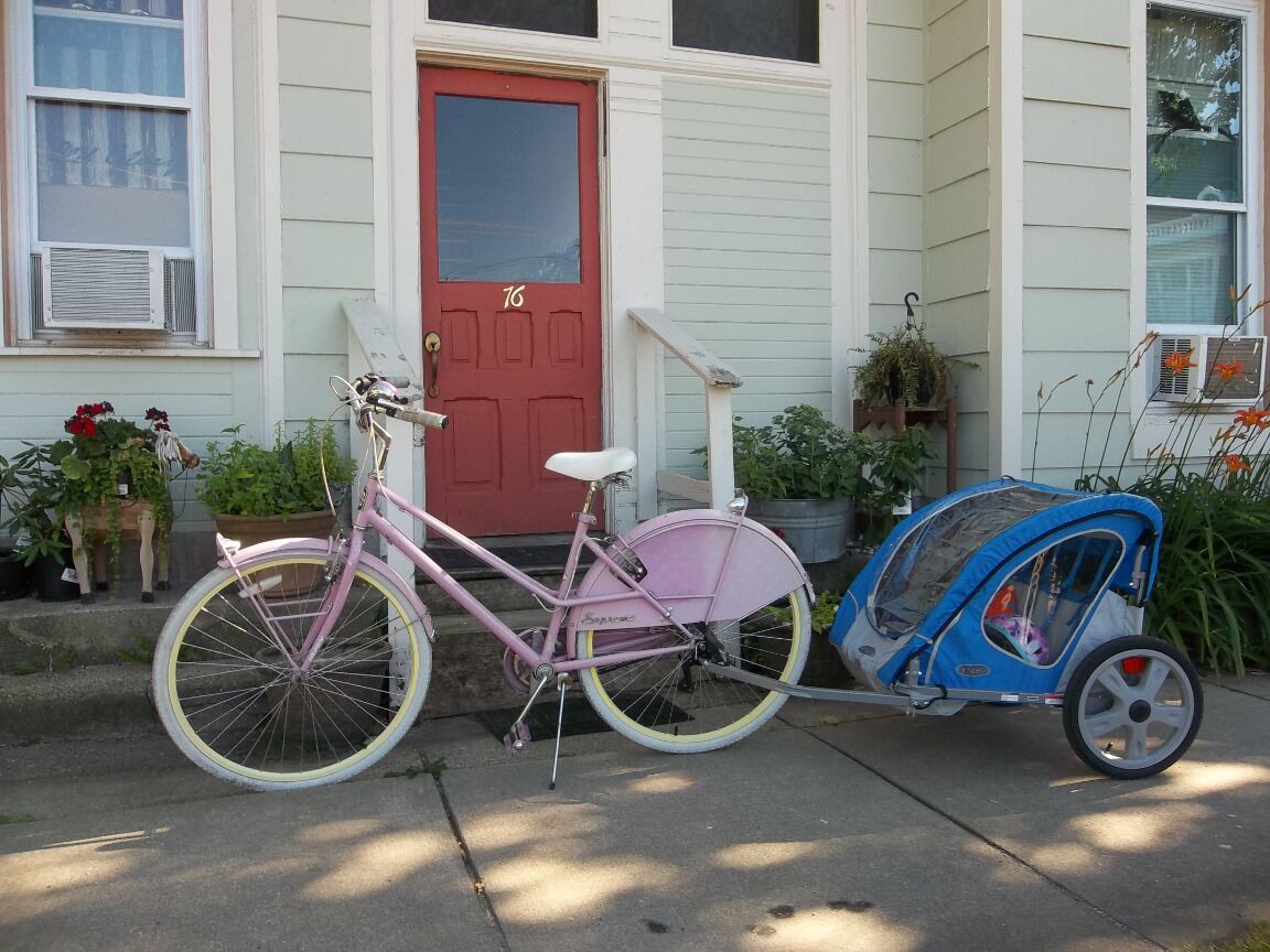 Een roze fiets met kinderzitje geparkeerd voor een huis met een rode deur en witte gevel.
