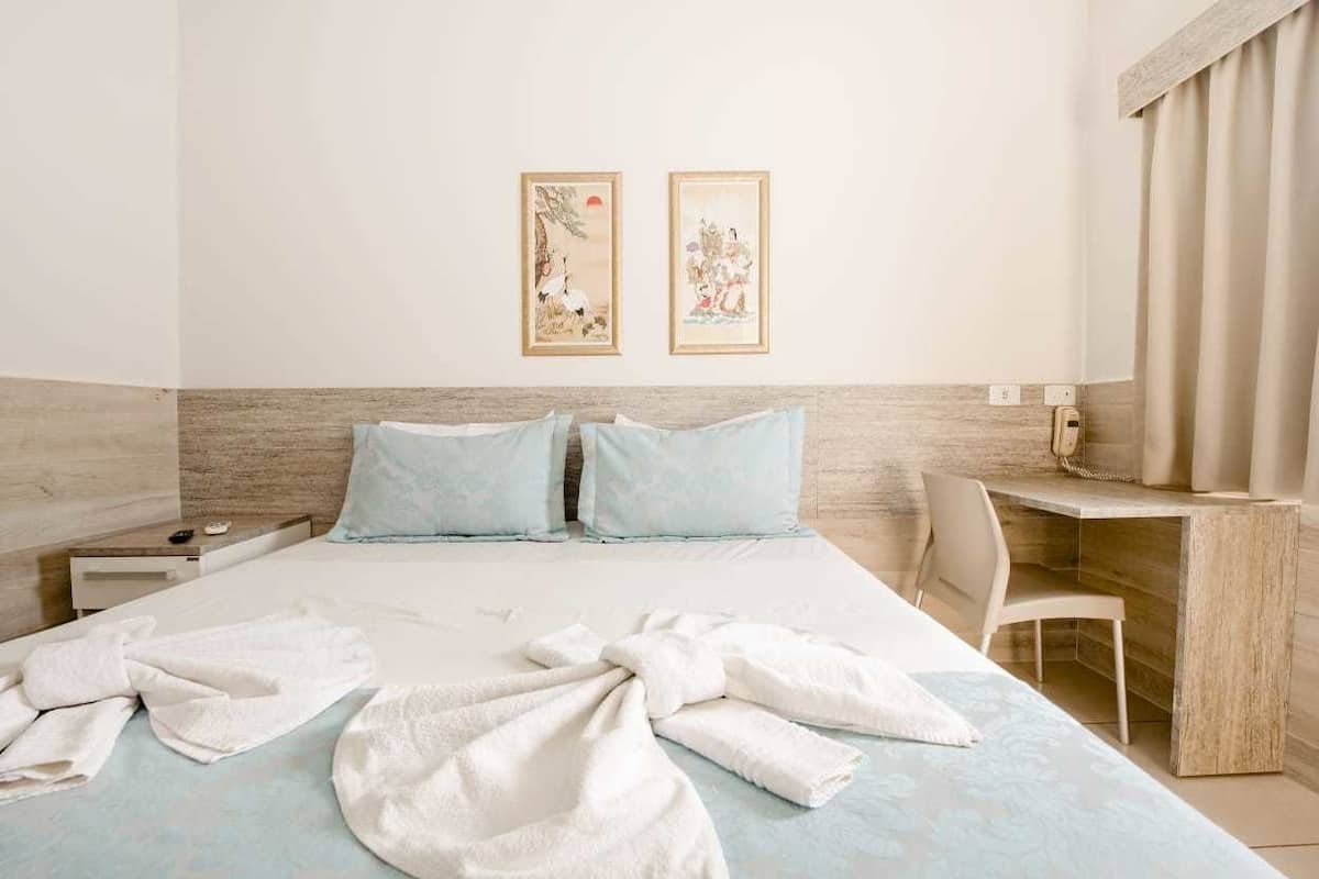 A hotel room with a bed, two pillows, a desk, a chair, and two framed artworks on the wall.