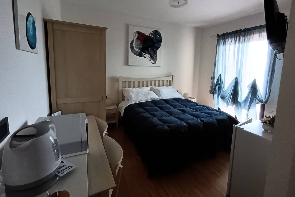 A bedroom with a bed, a desk with a kettle, a chair, a wardrobe, and a painting on the wall.