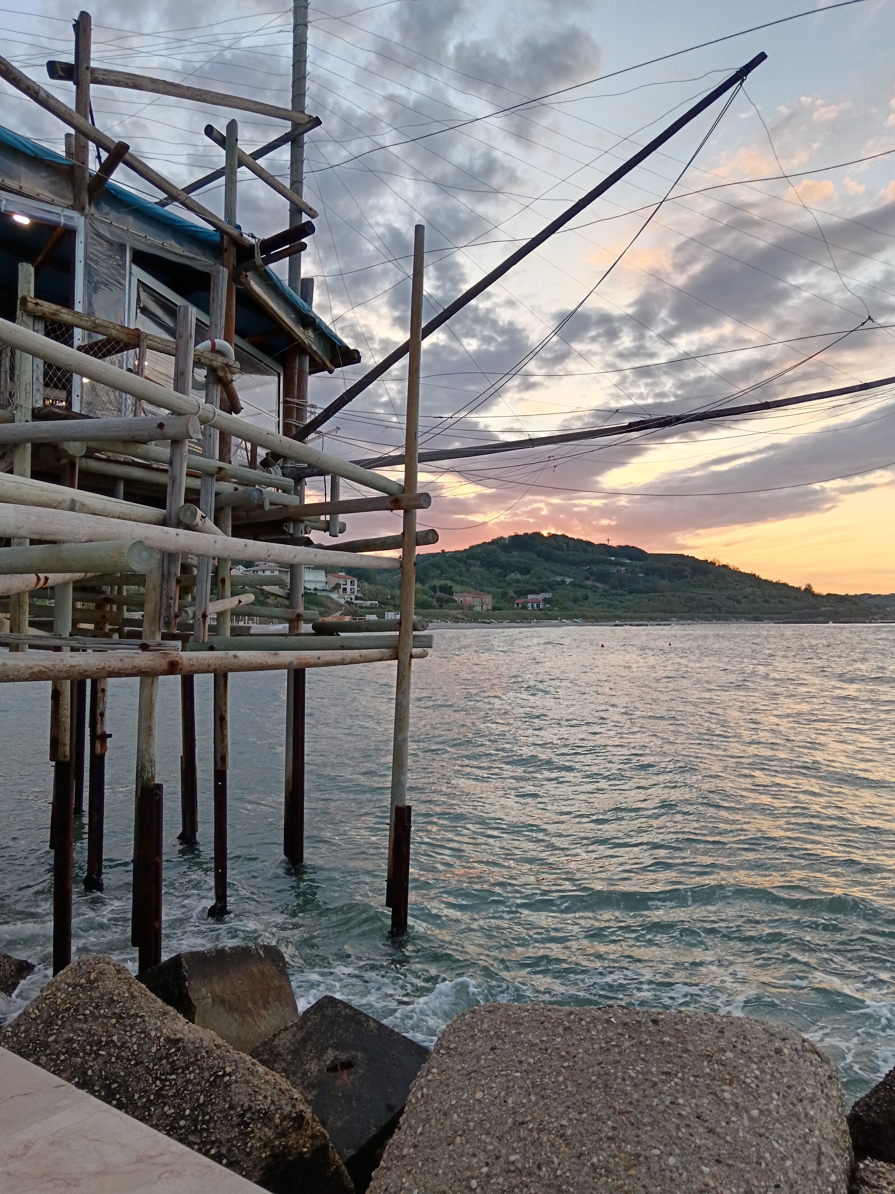 Un muelle con un edificio sobre pilotes, postes de madera y vistas al agua y colinas distantes.