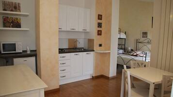 Cocina compacta con muebles blancos, un microondas y un lavabo. Hay una pequeña zona de comedor con una mesa y sillas.