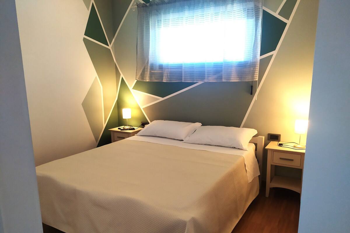 A neatly made bed with a white comforter and pillows, two bedside tables with lamps, a geometric-patterned wall, and a window with a sheer curtain.