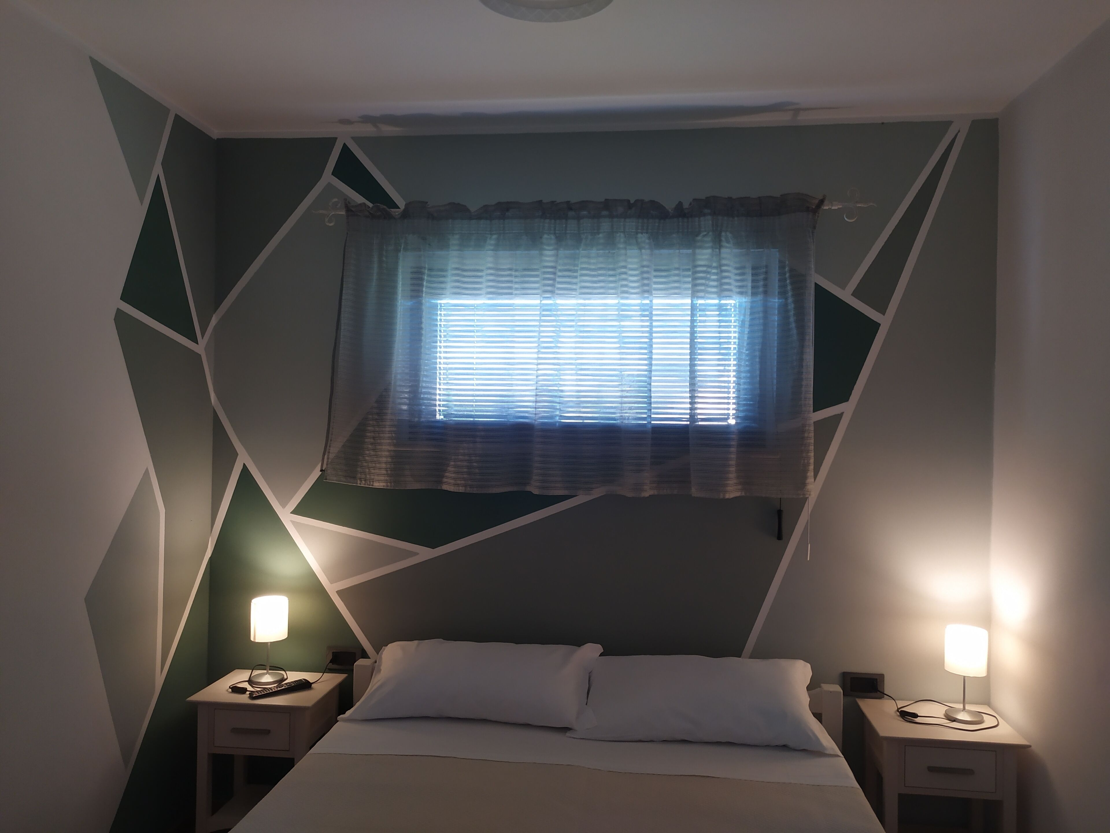 A bedroom with a bed, two bedside tables, a window with curtains, and a geometric wall design.
