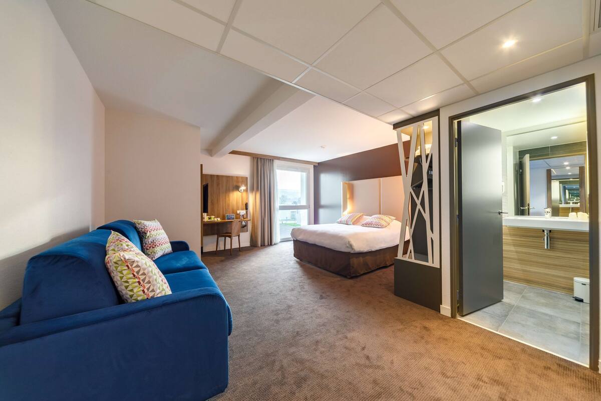 A modern hotel room with a blue sofa, a bed, a desk, and a bathroom visible through an open door.