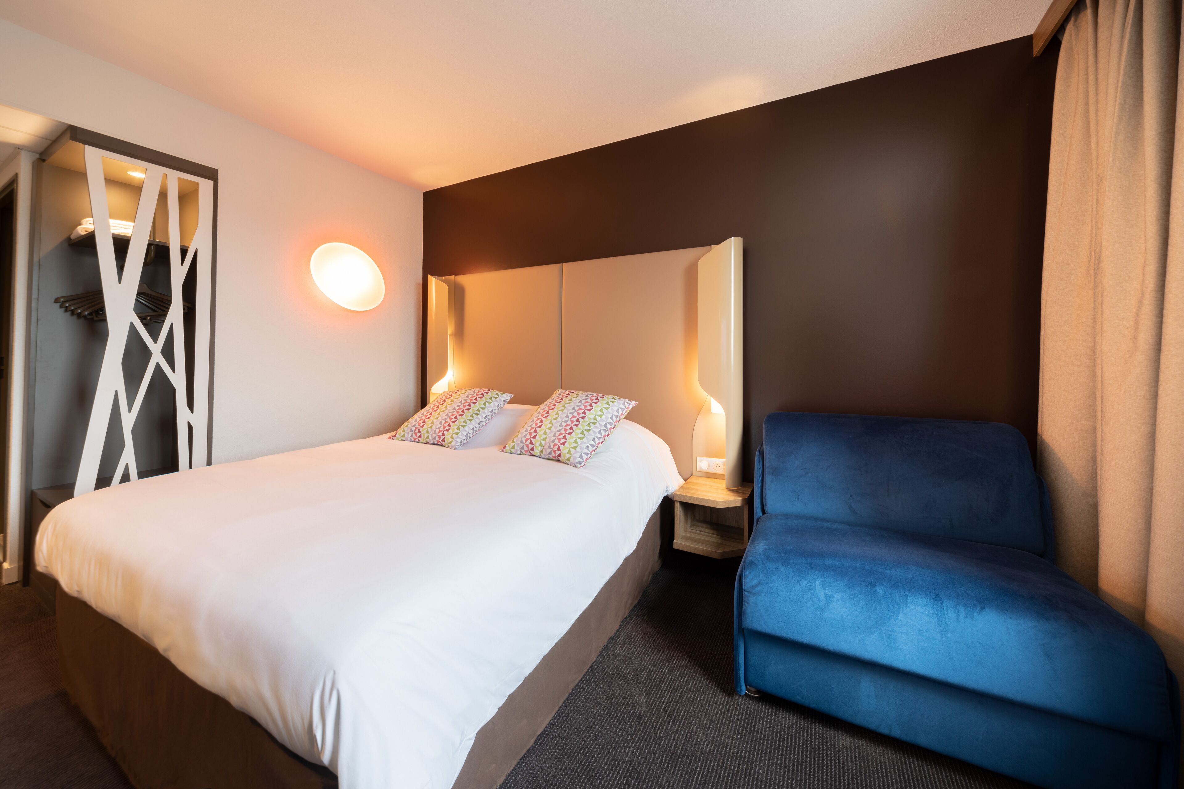 A hotel room with a large bed, a blue armchair, a wall-mounted light, and a decorative white partition.