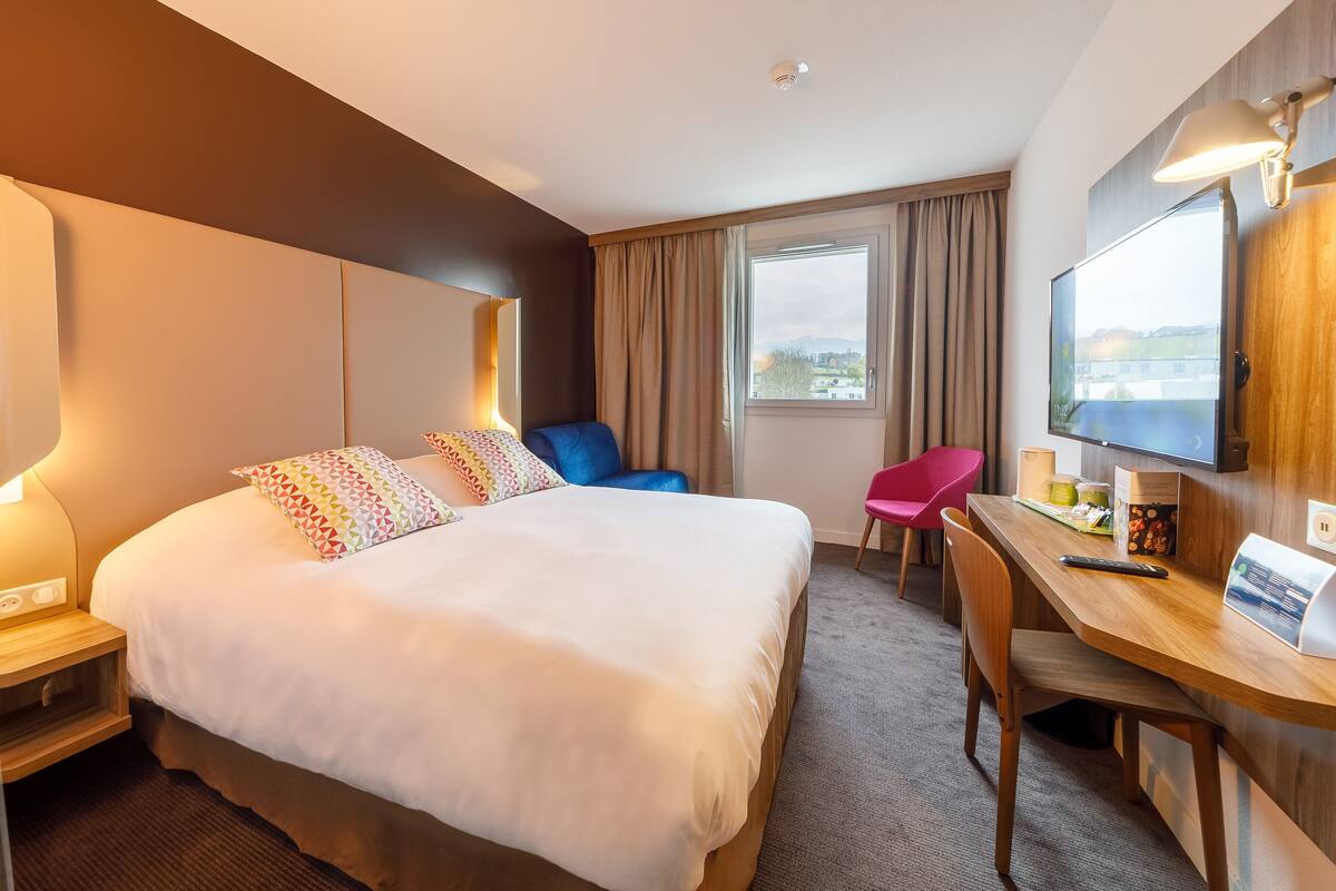 A hotel room with a large bed, a desk with a chair, a television, and a window with curtains.