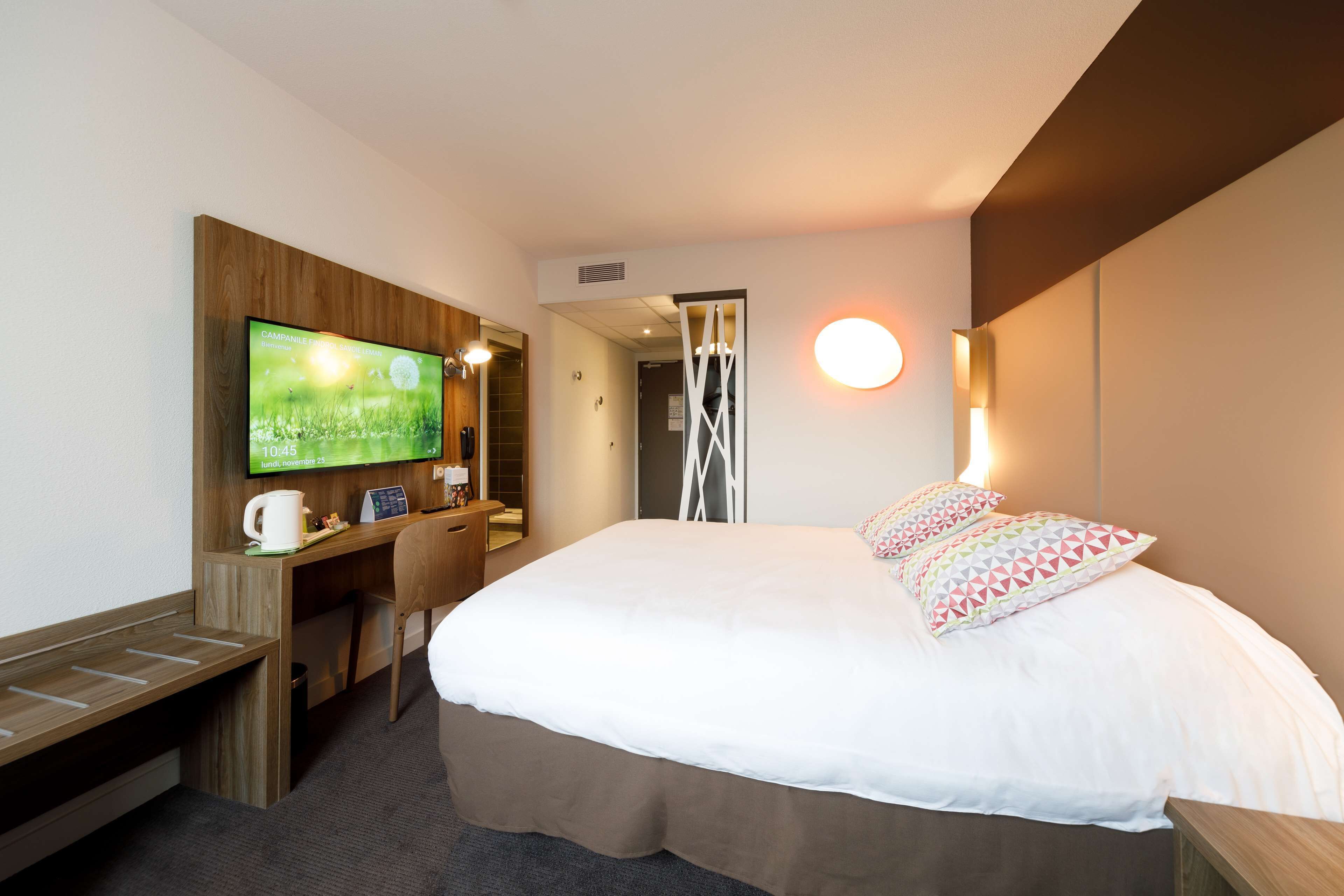 Ett hotellrum med en säng, ett skrivbord med en stol, en tv och en väggmonterad lampa.