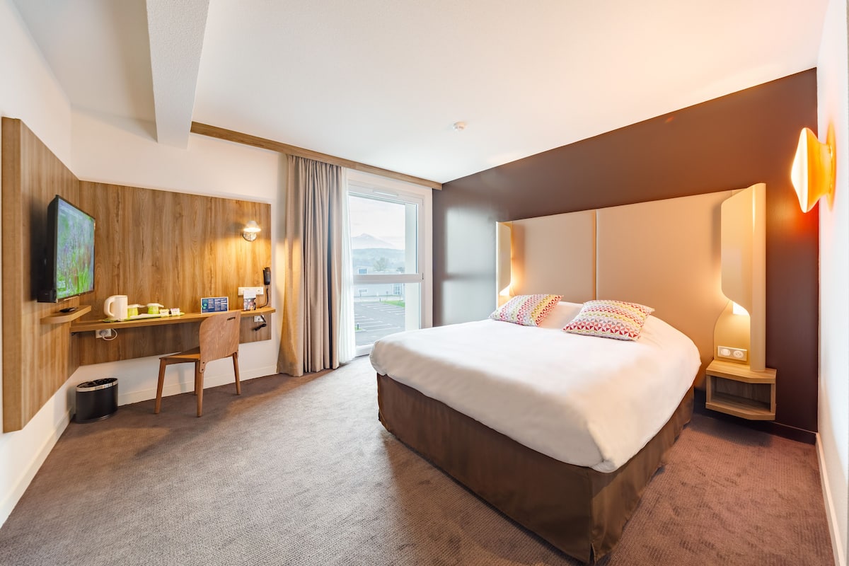 A hotel room with a large bed, a desk with a chair, a television, and a window with curtains.