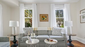 Ein Wohnzimmer mit einem grauen Sofa, zwei weißen Nachttischen mit Lampen, einem gemusterten Teppich und einem Fenster mit Vorhängen.