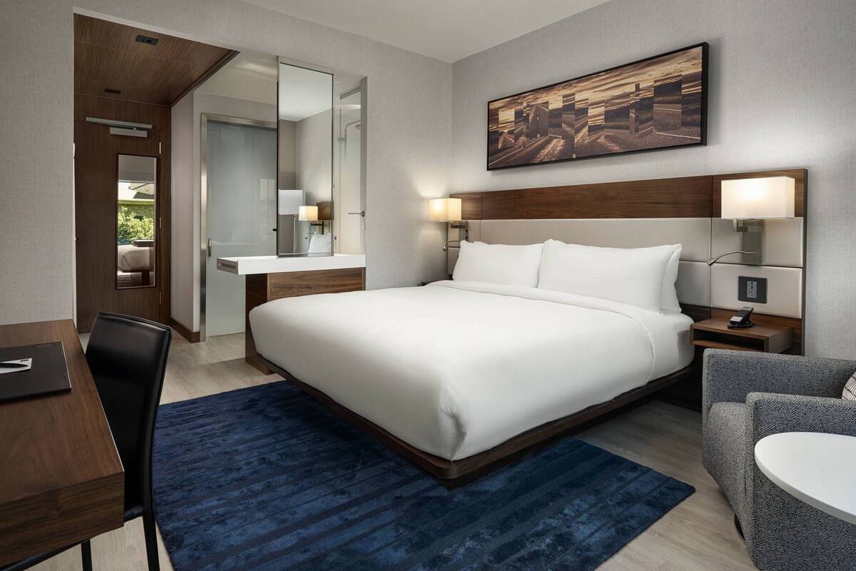 A hotel room with a large bed, a desk, a chair, a small table, a mirror, and a painting on the wall.