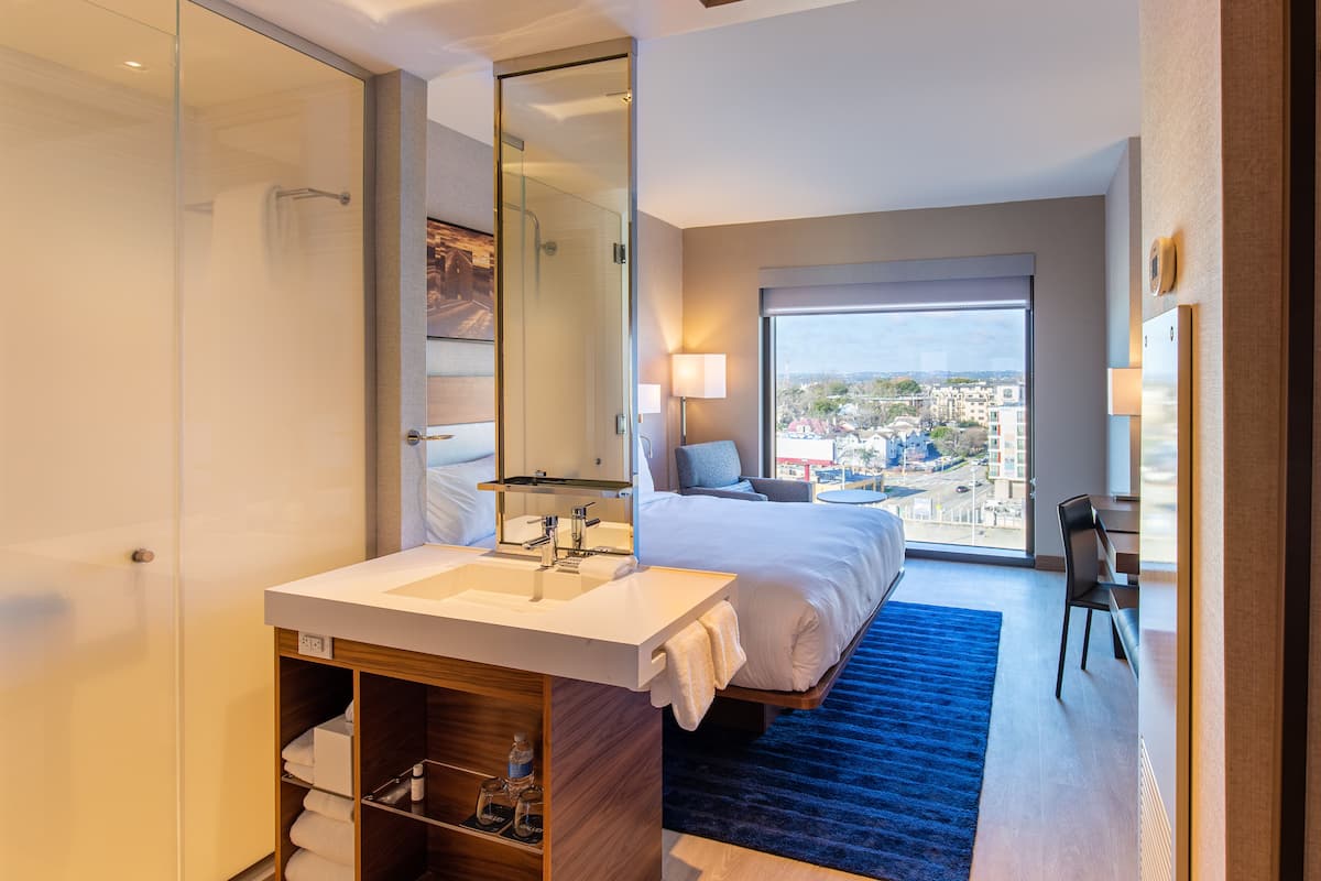 A hotel room with a large bed, a bathroom with a glass shower enclosure, a white sink, and a city view through the window.