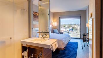 A hotel room with a large bed, a bathroom with a glass shower enclosure, a white sink, and a city view through the window.