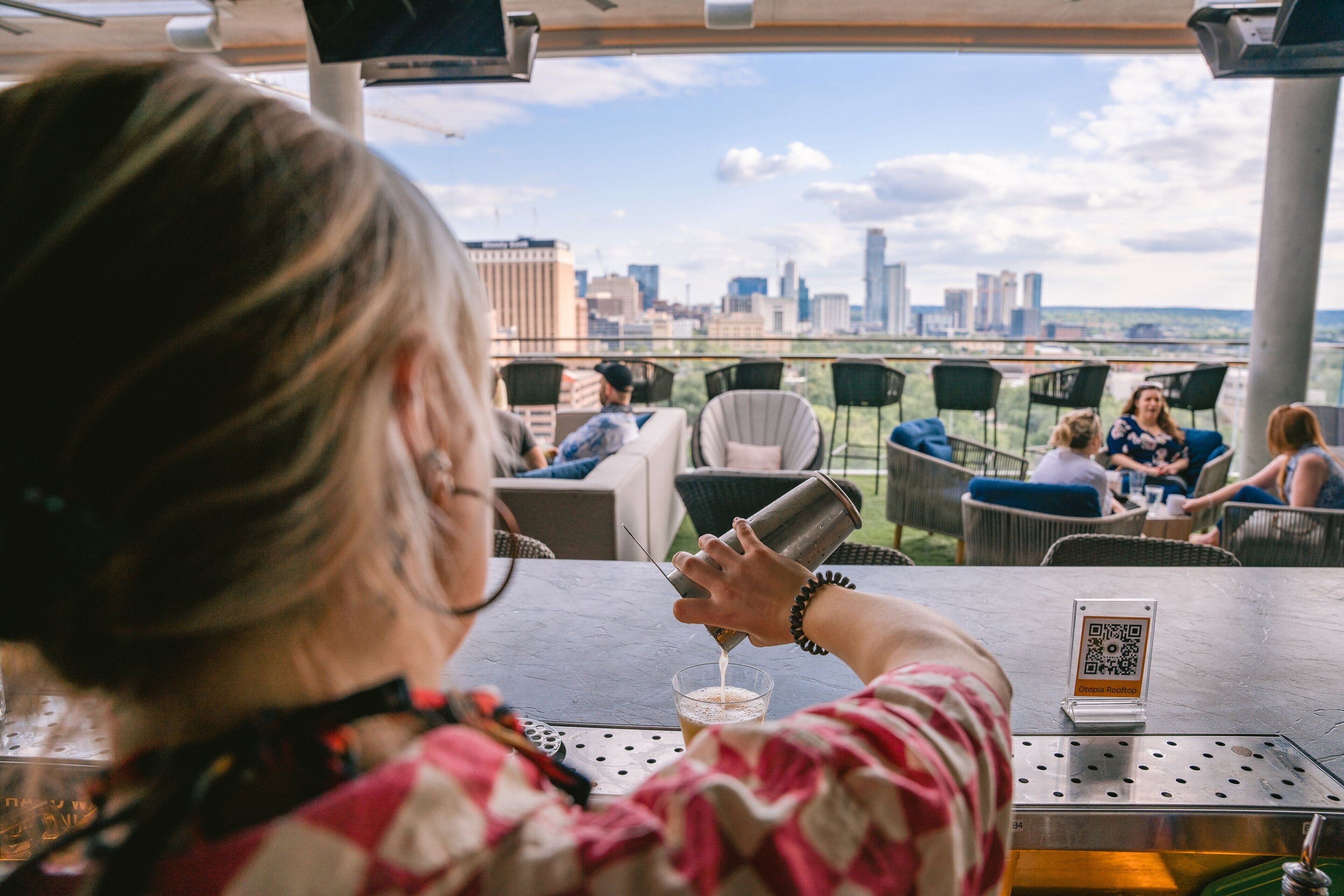 A person pouring a drink at a bar with a city view, outdoor seating, and a QR code on the table.