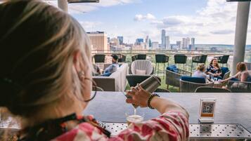A person pouring a drink at a bar with a city view, outdoor seating, and a QR code on the table.