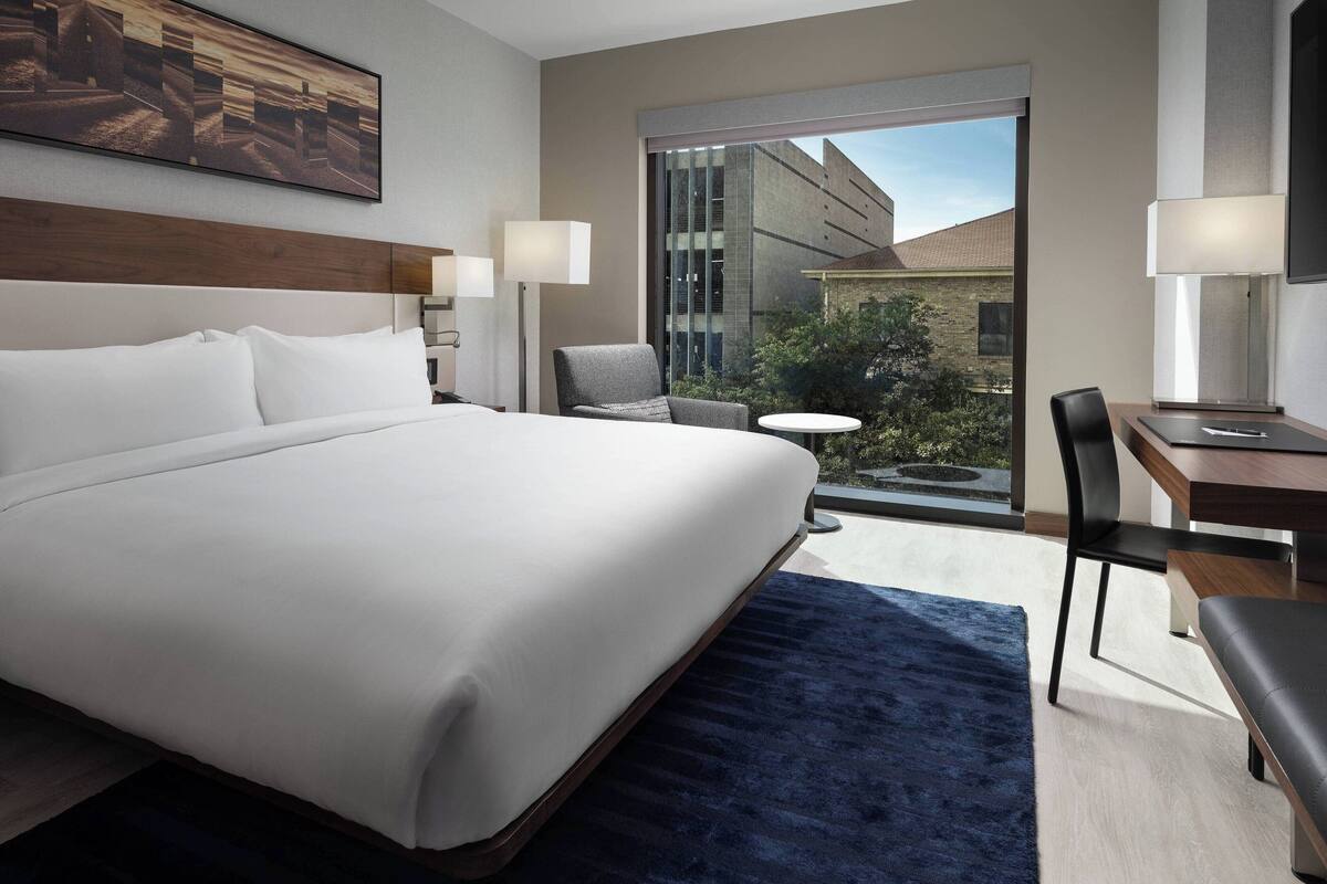 A hotel room with a large bed, a desk, a chair, a small table, and a view of the outside.