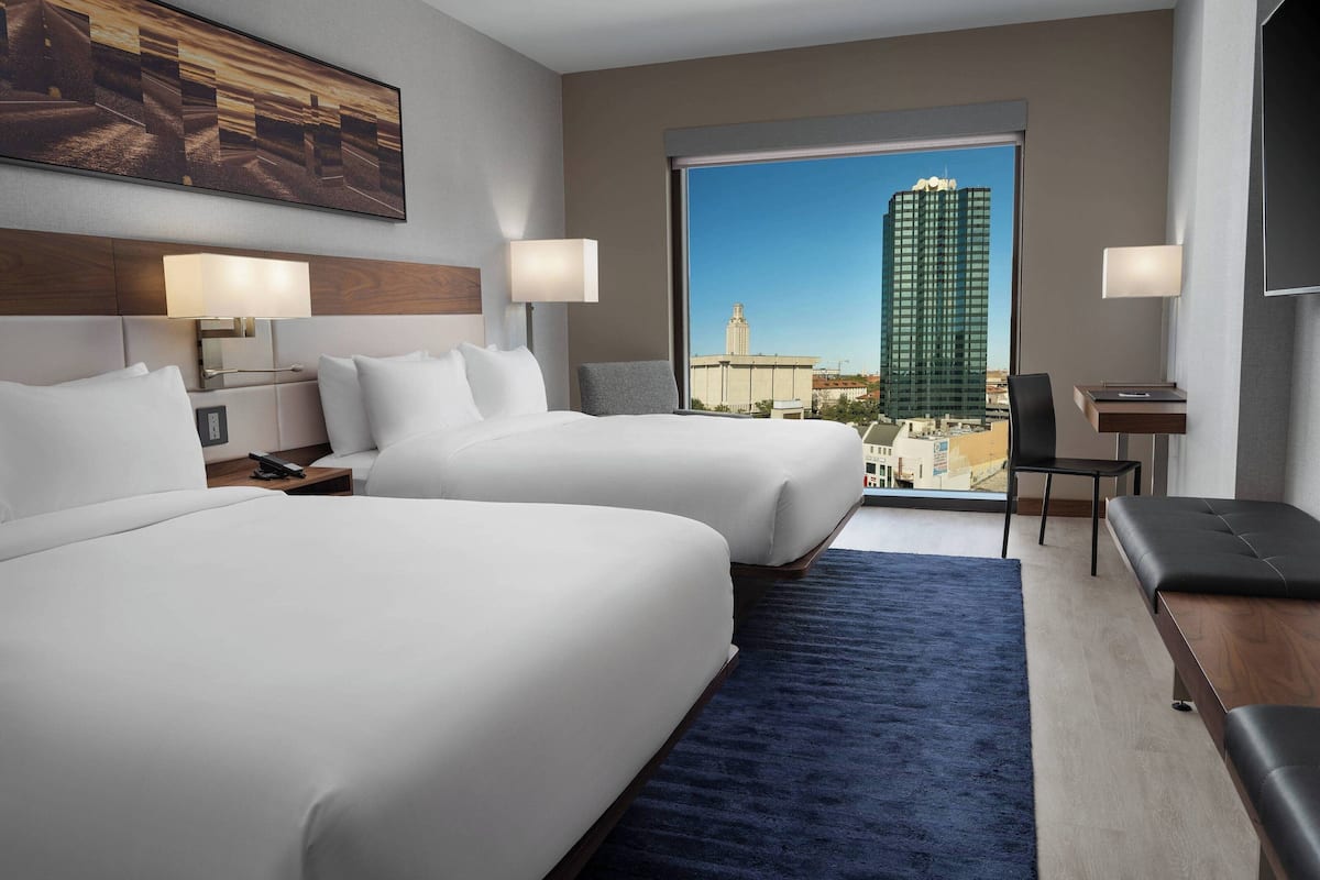 A hotel room with two beds, a desk, a chair, and a large window with a city view.