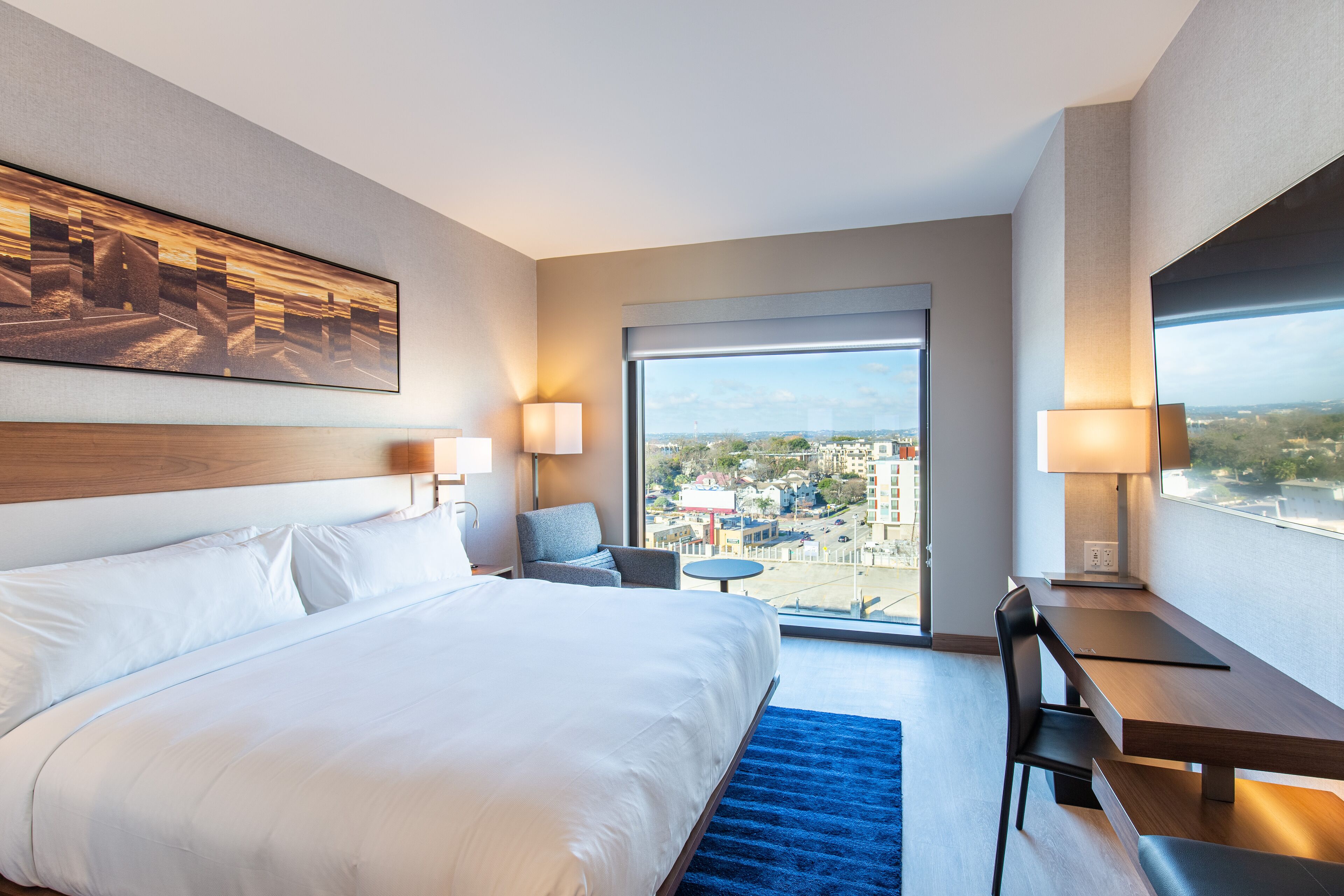 Hotelkamer met een groot bed, een bureau, twee stoelen, een kleine tafel en een groot raam met uitzicht op de stad.