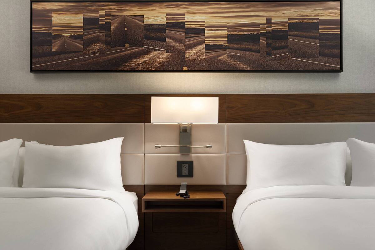 A hotel room with two beds, a nightstand with a lamp, a wall-mounted artwork, and a wooden headboard.