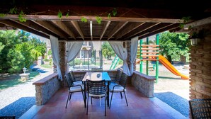 A covered patio with a table and chairs, overlooking a playground with a slide. - Apartment Sanic Rab 2 (Lopar)