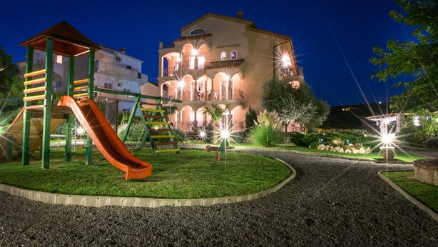 A playground with a slide and climbing structures, surrounded by a well-maintained garden and a large building with multiple balconies and arches in the background. - Apartment Sanic Rab 2 (Lopar)