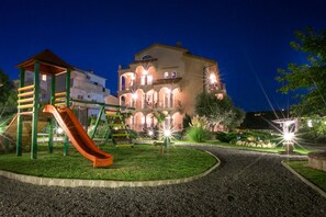 Un'area giochi con scivolo e strutture per arrampicarsi, circondata da un giardino curato e un edificio con numerosi balconi e arcate sullo sfondo.