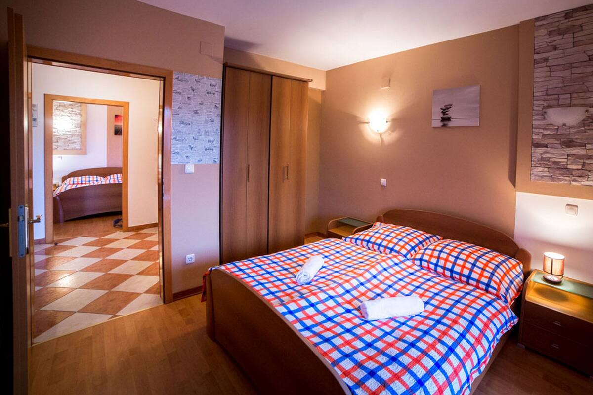 A hotel room with a checkered floor, a bed with a plaid bedspread, a wooden wardrobe, a nightstand with a lamp, and a wall-mounted light fixture.