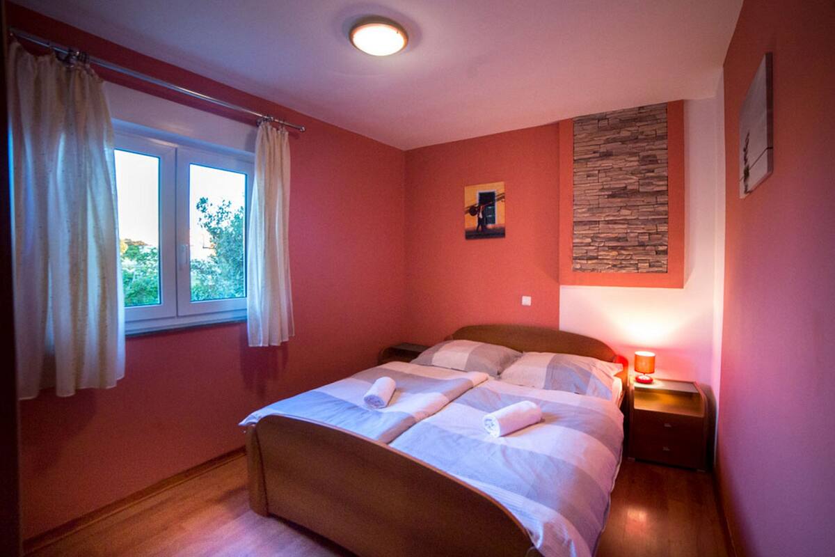 A bedroom with two beds, a window with curtains, a nightstand with a lamp, and a wall-mounted artwork.