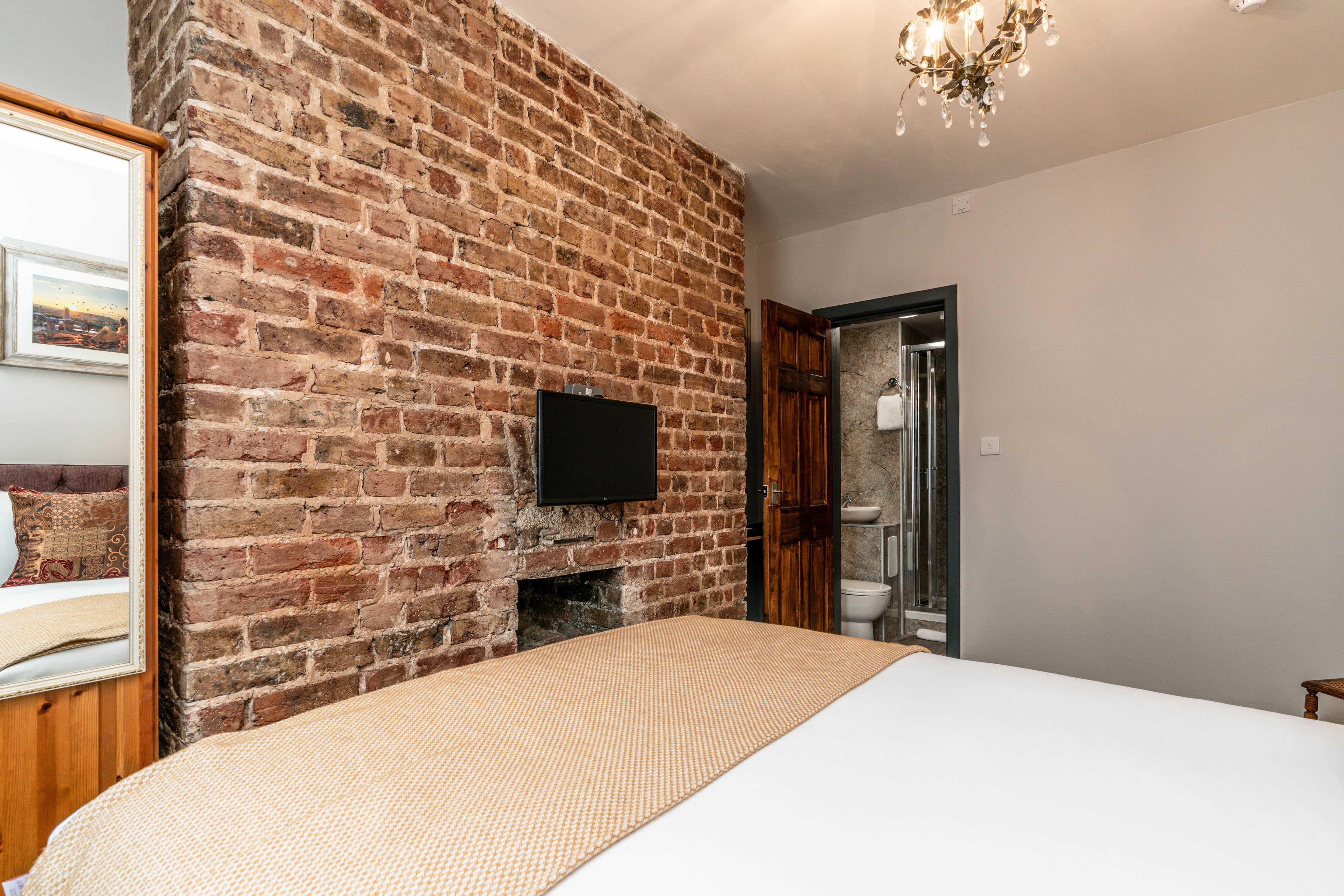 Une chambre avec un mur en briques, un téléviseur à écran plat, un lit et une salle de bain visible à travers une porte ouverte.