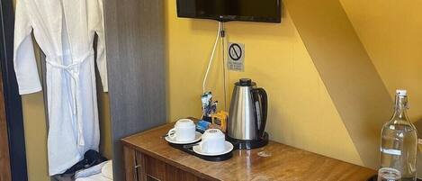 A hotel room with a wooden vanity, a flat-screen TV mounted on the wall, a robe hanging on a rack, and a small table with a coffee pot and cups.