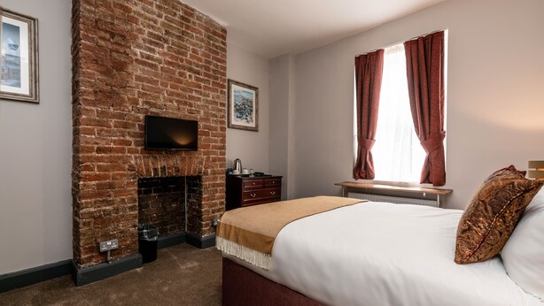 A bedroom with a brick fireplace, a bed with white bedding, a window with curtains, and a flat-screen TV mounted on the wall.