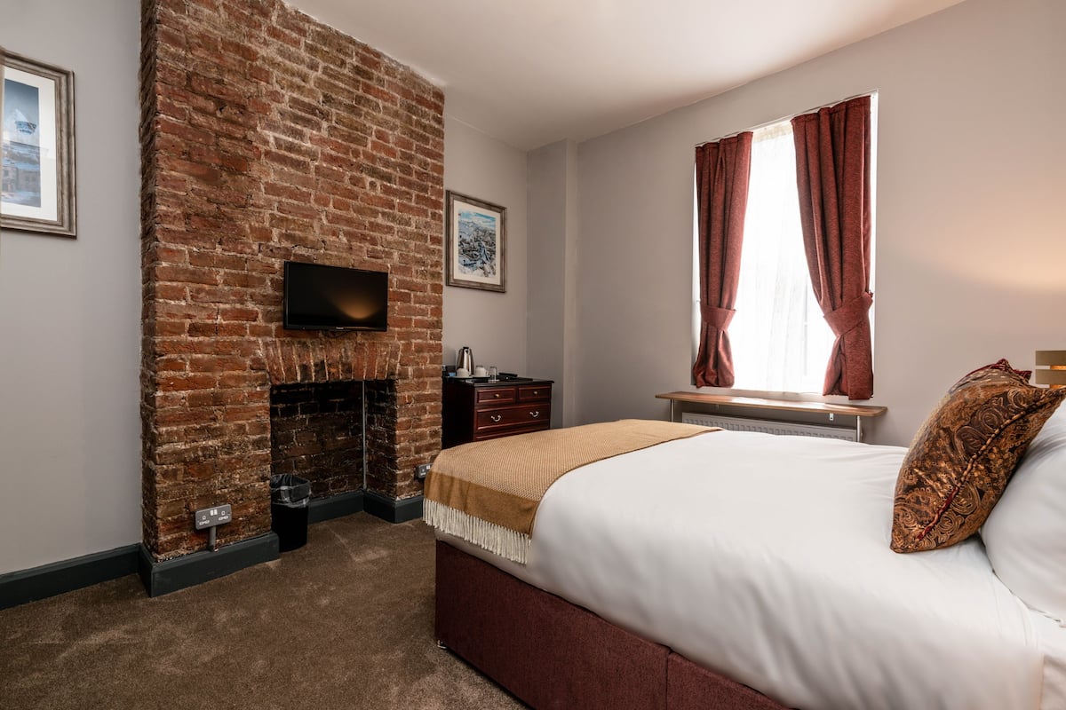 A bedroom with a brick fireplace, a bed with white bedding, a window with curtains, and a flat-screen TV mounted on the wall.