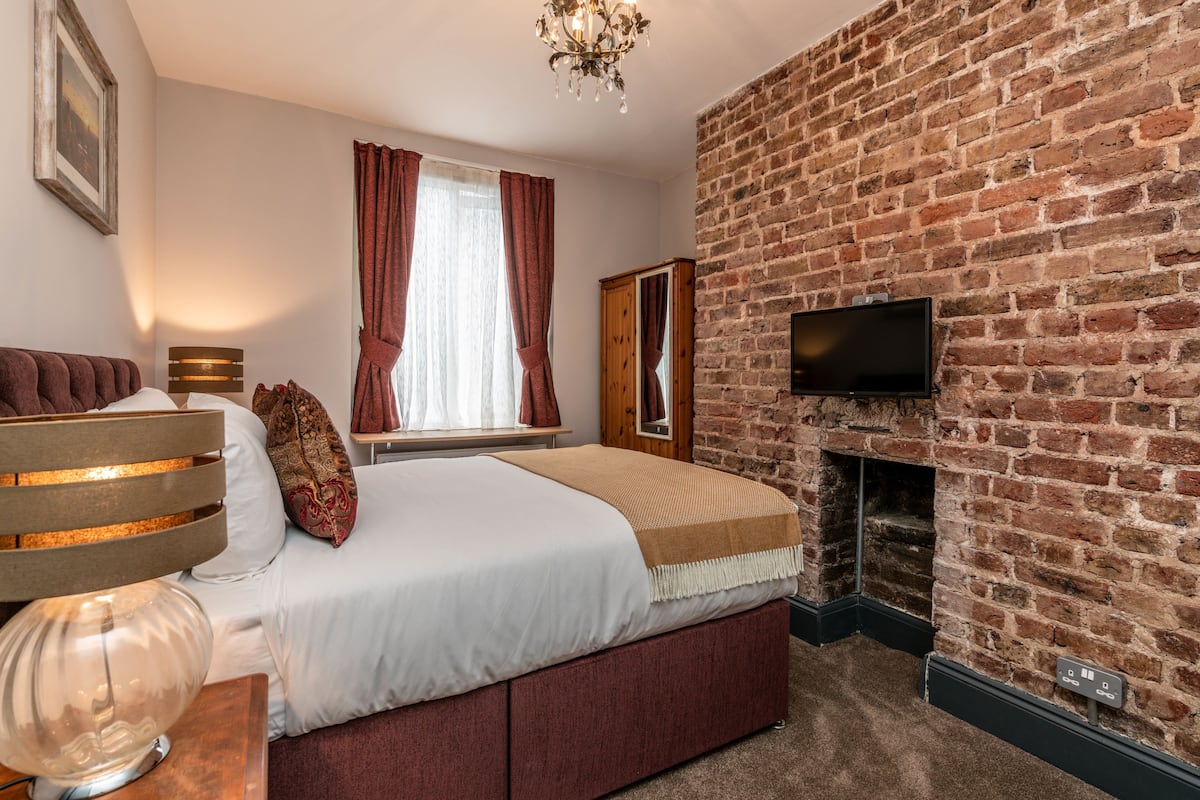 A bedroom with a brick wall, a fireplace, a bed, a TV, and a window with curtains.
