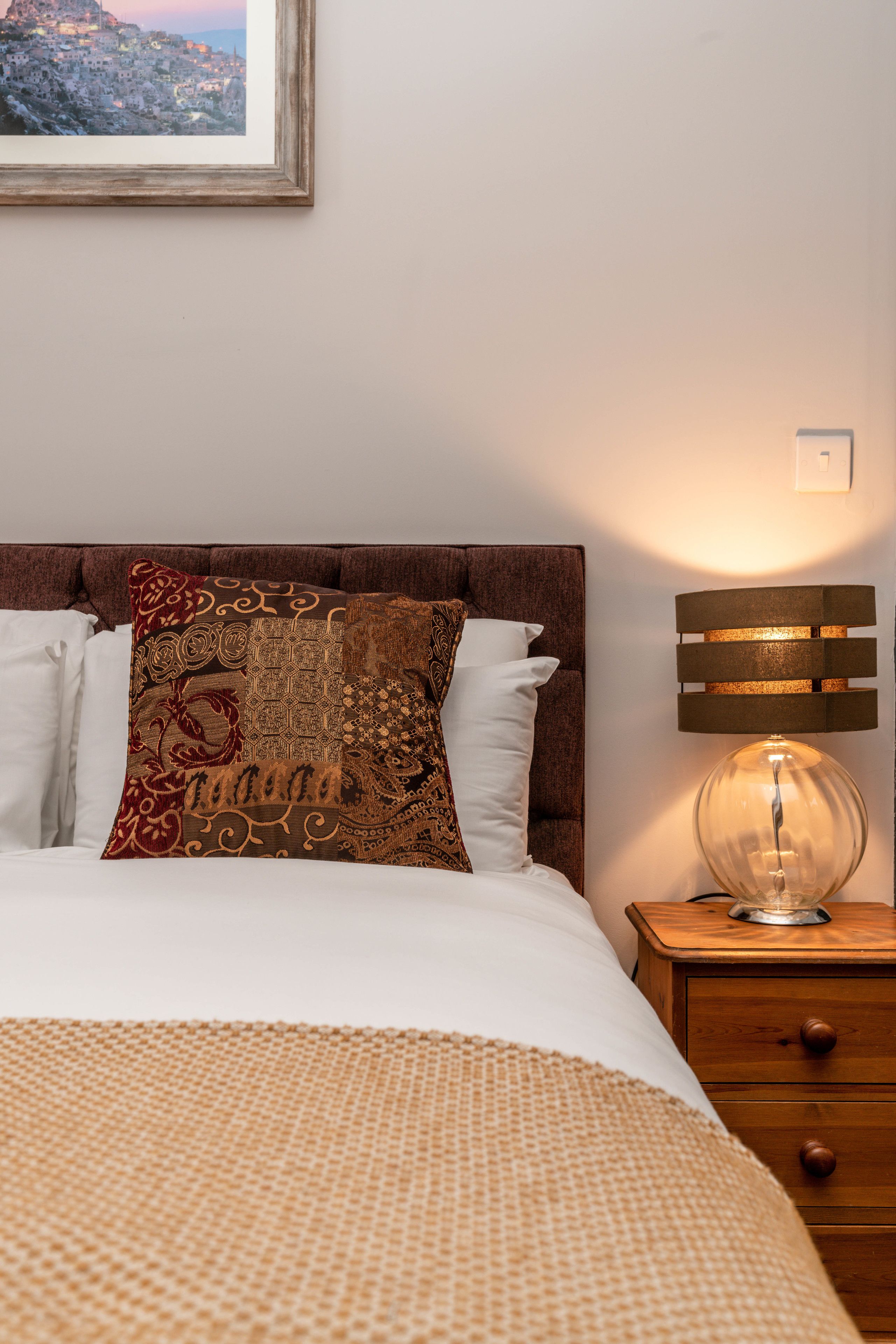 A neatly made bed with patterned pillows, a wooden nightstand with a lamp, and a framed picture on the wall.