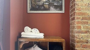 A room with a wooden shelf holding rolled towels and a white robe hanging. A framed picture of a town is on the wall above the shelf.