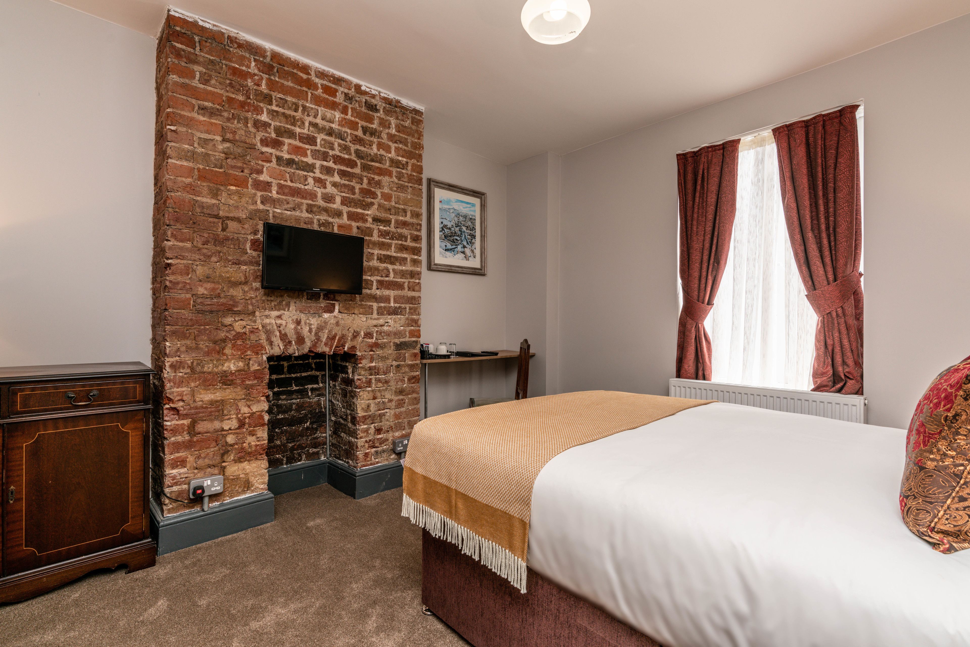 Une chambre d’hôtel avec une cheminée en briques, un lit recouvert d’une couverture blanche et marron, une commode en bois, un téléviseur à écran plat fixé au mur et une fenêtre avec des rideaux rouges.