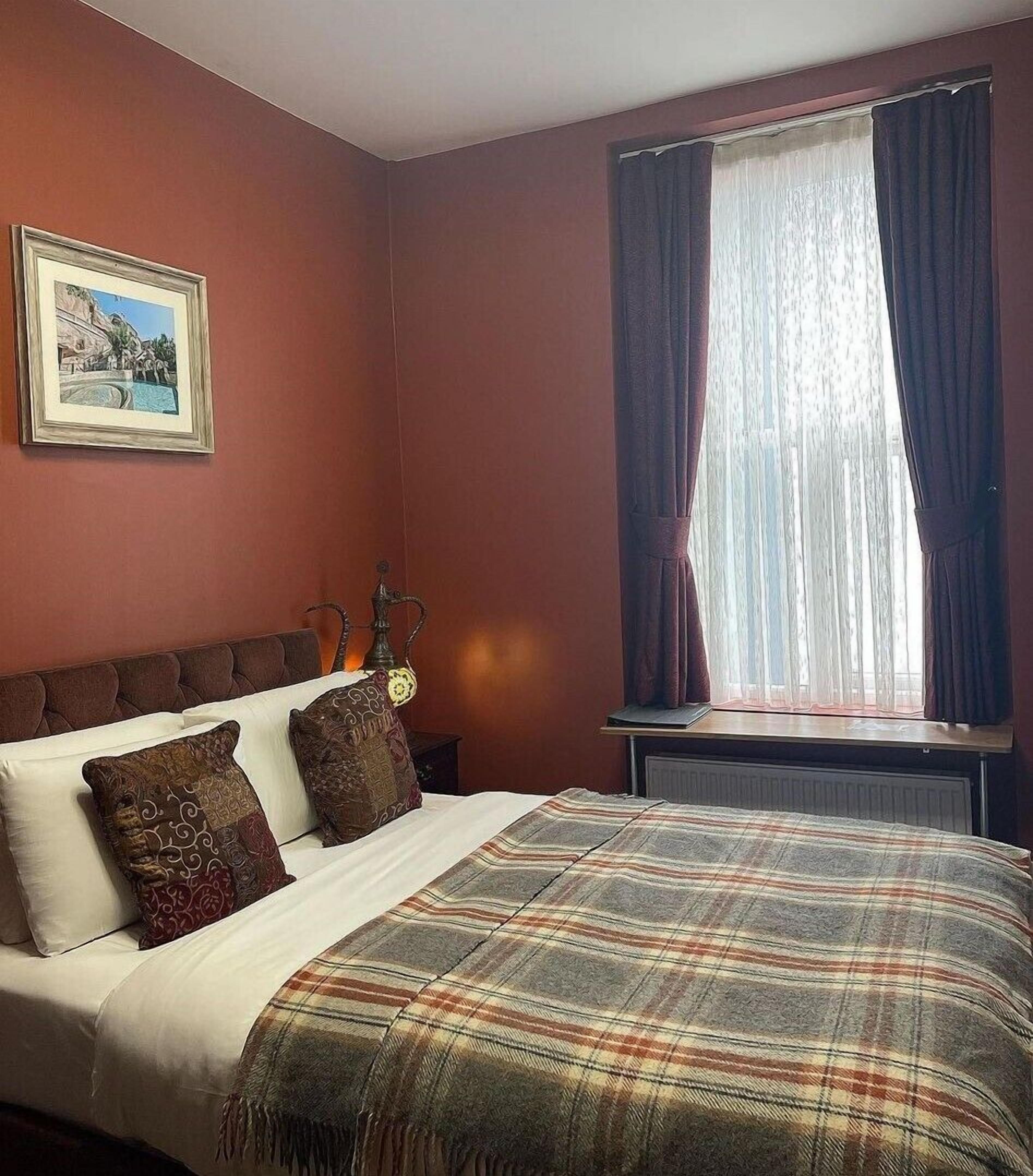 Habitación de hotel con cama, mesita de noche, lámpara, ventana con cortinas y un cuadro en la pared.