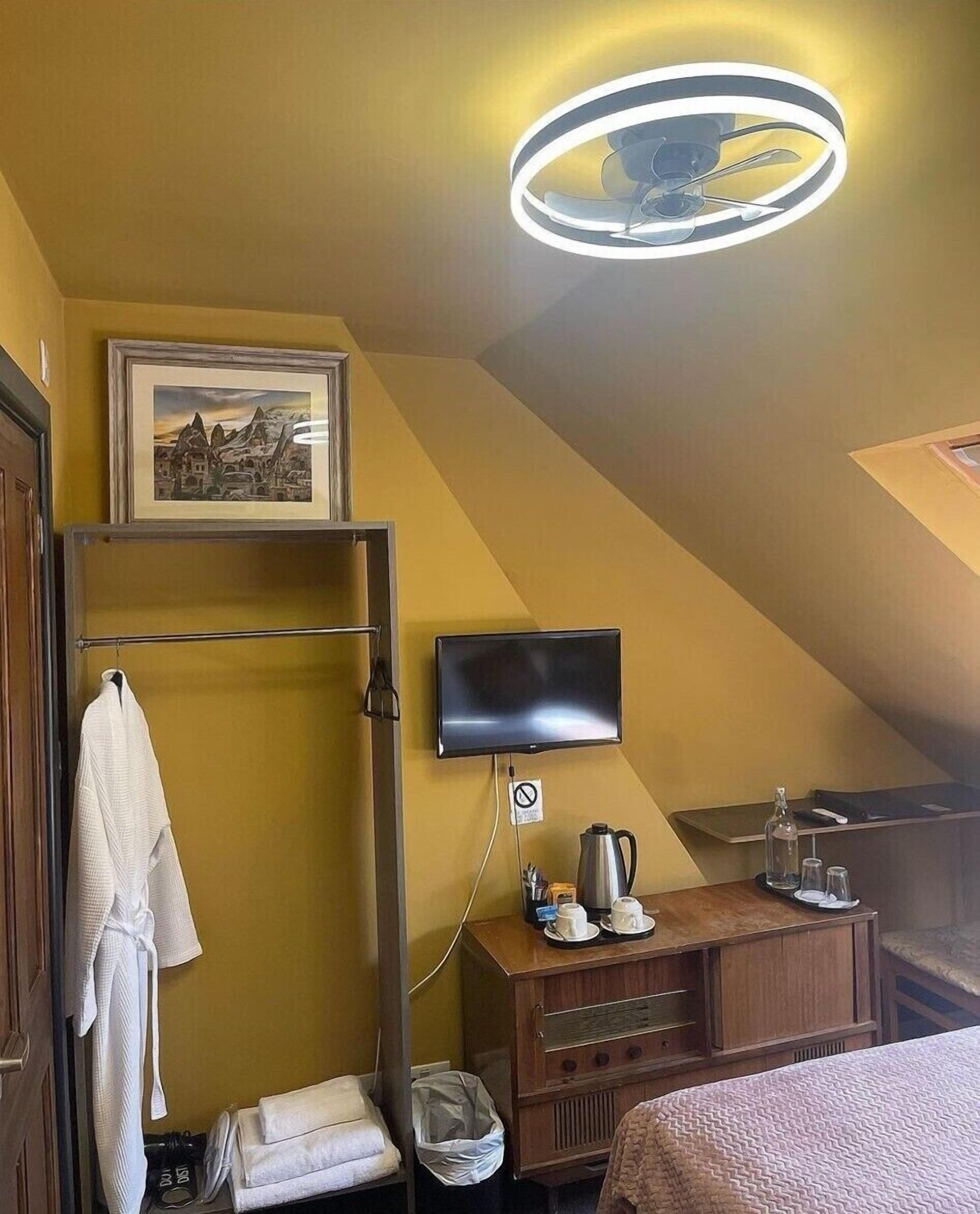 A hotel room with a bed, a TV mounted on the wall, a framed picture above the bed, a towel rack, a wooden dresser, and a small table with a coffee pot and cups.