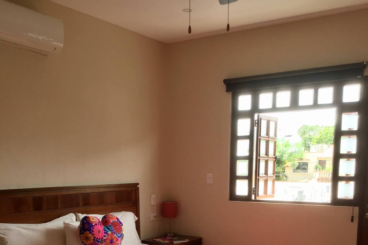 A bedroom with a bed, a nightstand, a red lamp, and a window with a view of buildings and greenery.