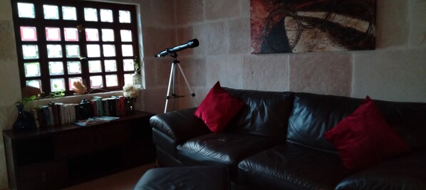 A living room with a black leather sofa, red cushions, a telescope on a tripod, and a window with a grid pattern.