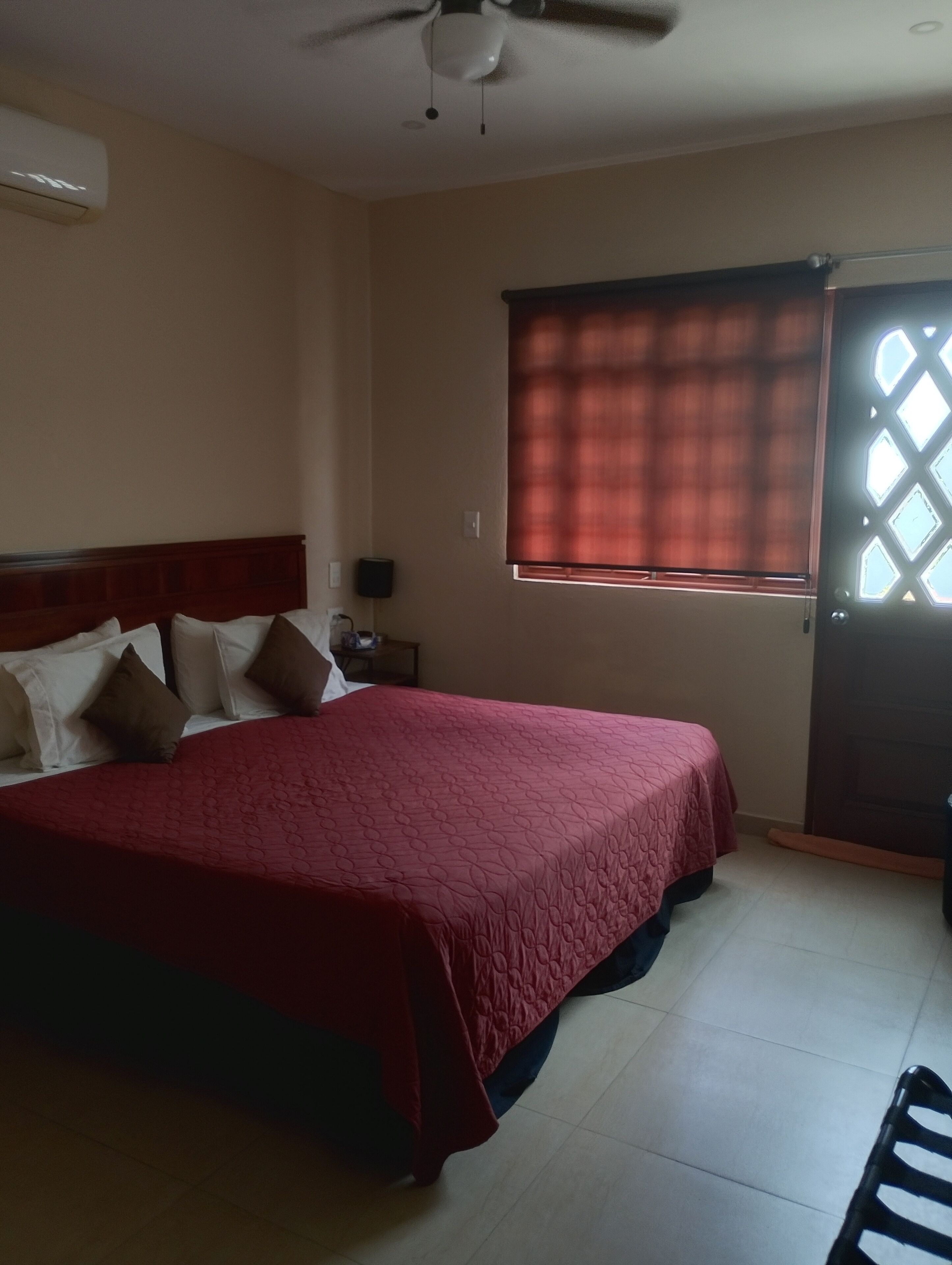 Quarto com cama, ventilador de teto, janela com persianas e porta com padrão geométrico.