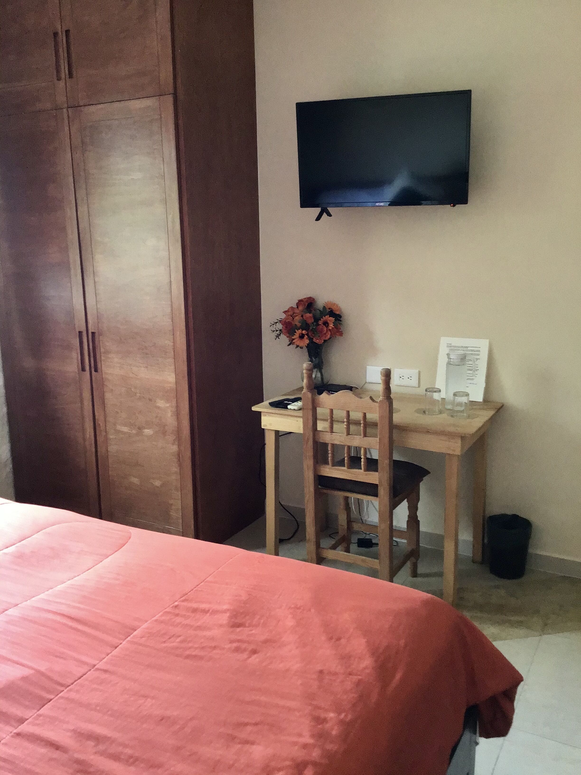 A bedroom with a bed, a wooden desk, a chair, a TV on the wall, and a vase with flowers.