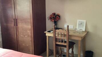 A bedroom with a bed, a wooden desk, a chair, a TV on the wall, and a vase with flowers.