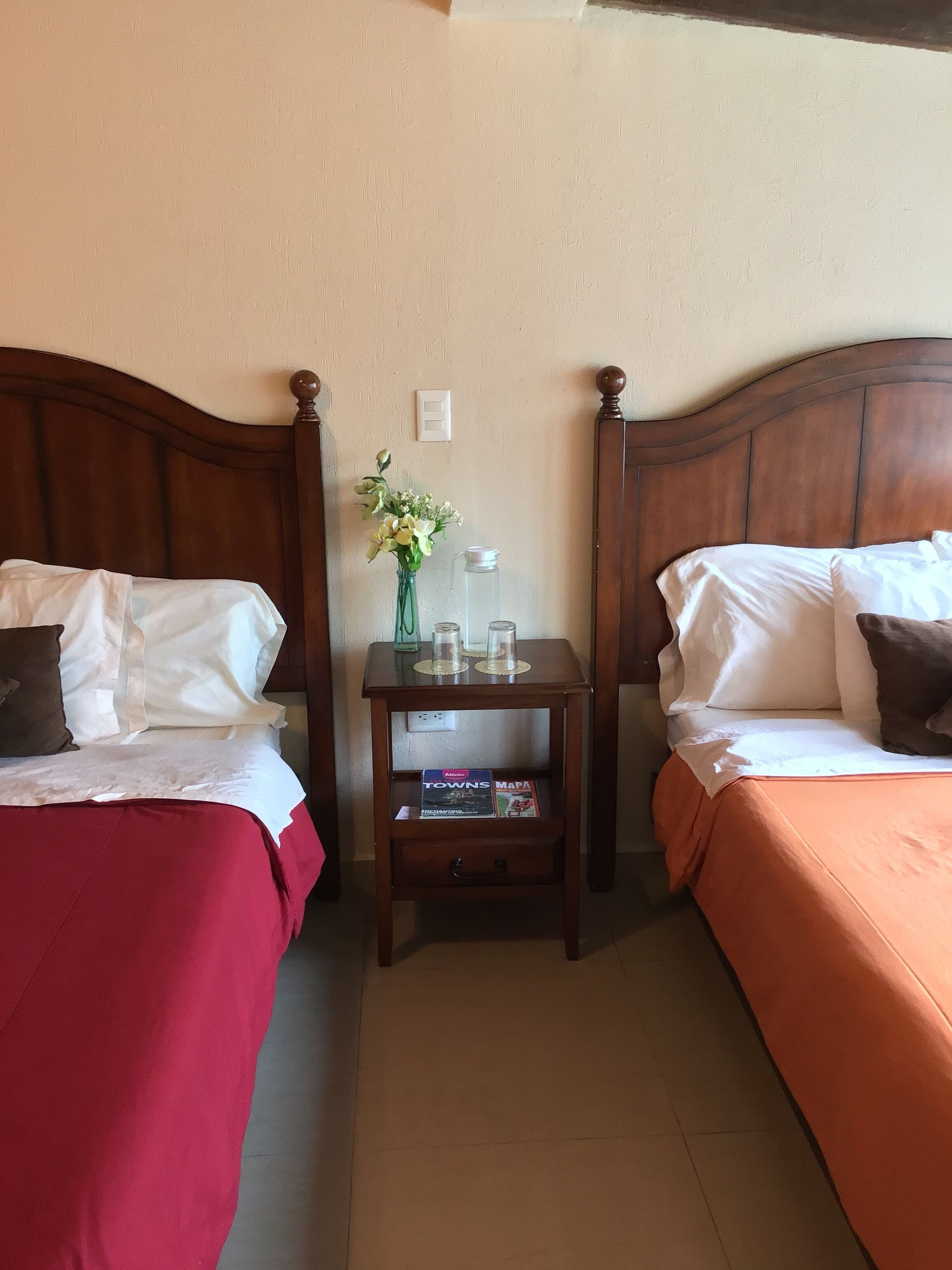 A hotel room with two single beds, a nightstand with a vase of flowers, and a book titled 'TOWNS MAPA'.