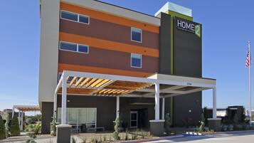 Un hotel moderno con una zona salotto esterna coperta, un cartello ben visibile con la scritta 'HOME 2' e un'asta con la bandiera americana.