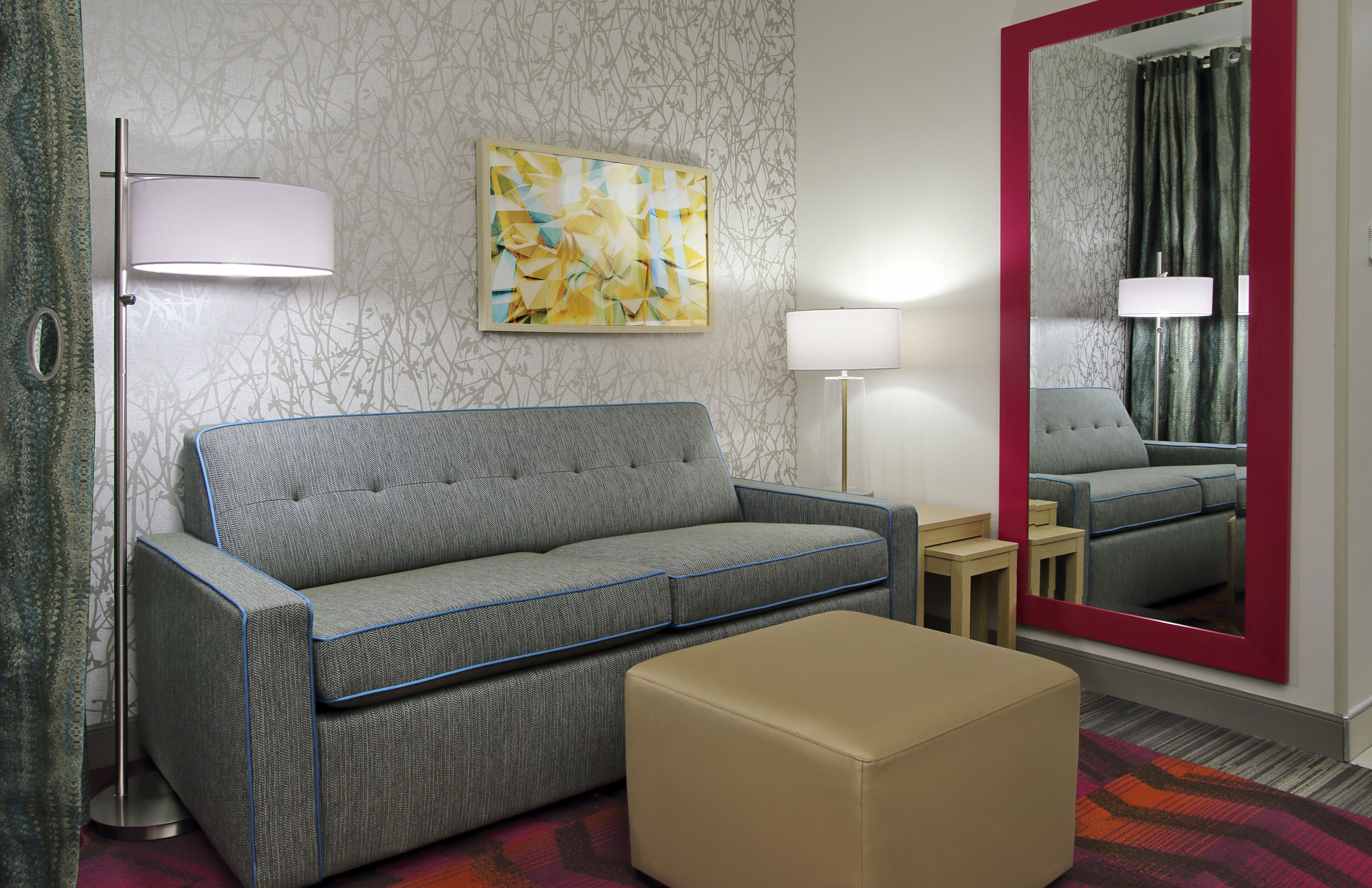 Moderni hotellihuone, jossa on harmaa sohva, beigenvärinen jalkarahi, lattiavalaisin ja suuri peili, jonka kehys on punainen.