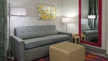 Moderni hotellihuone, jossa on harmaa sohva, beigenvärinen jalkarahi, lattiavalaisin ja suuri peili, jonka kehys on punainen.