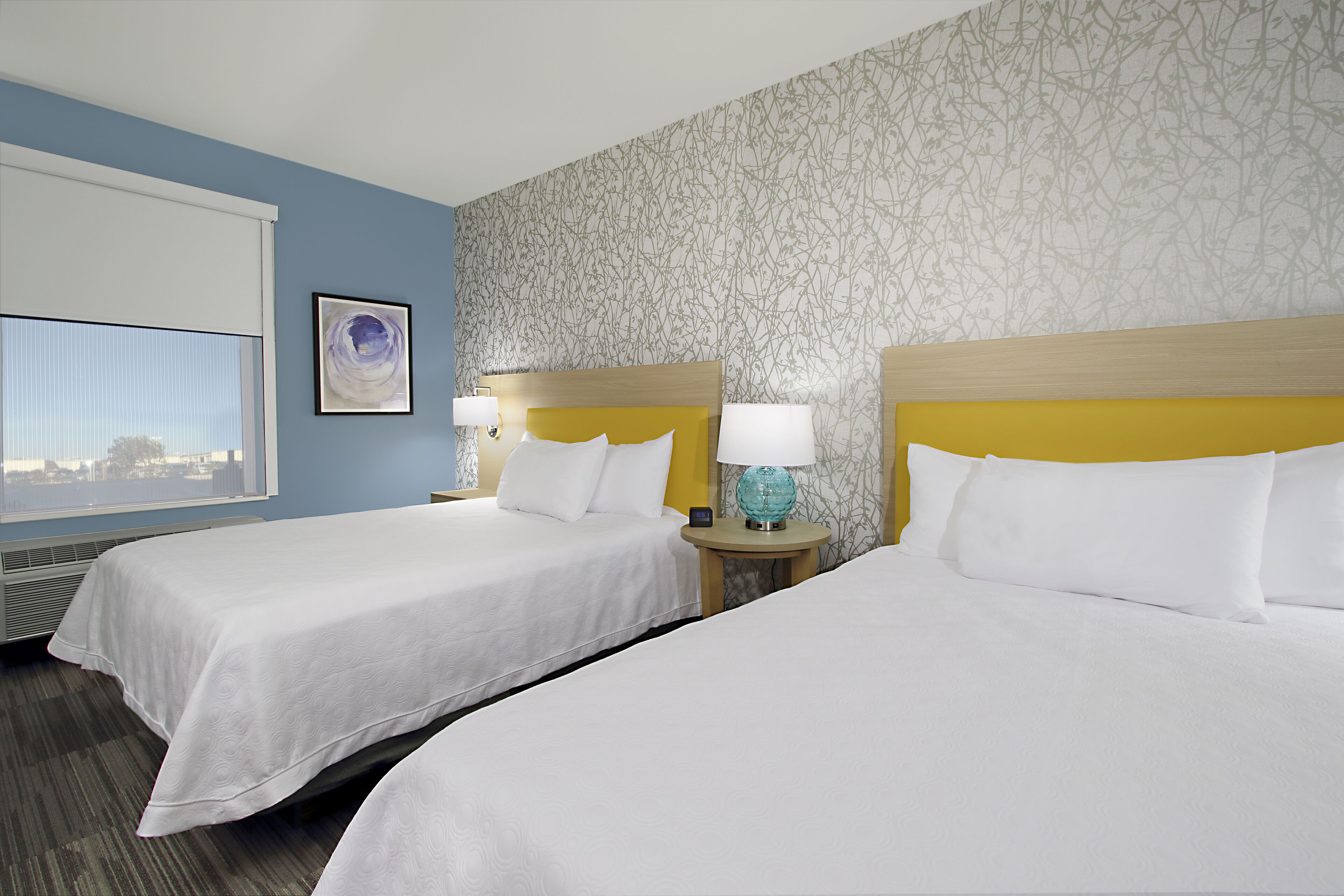 A hotel room with two beds, a window with blinds, a wall with a floral pattern, and a nightstand with a lamp.