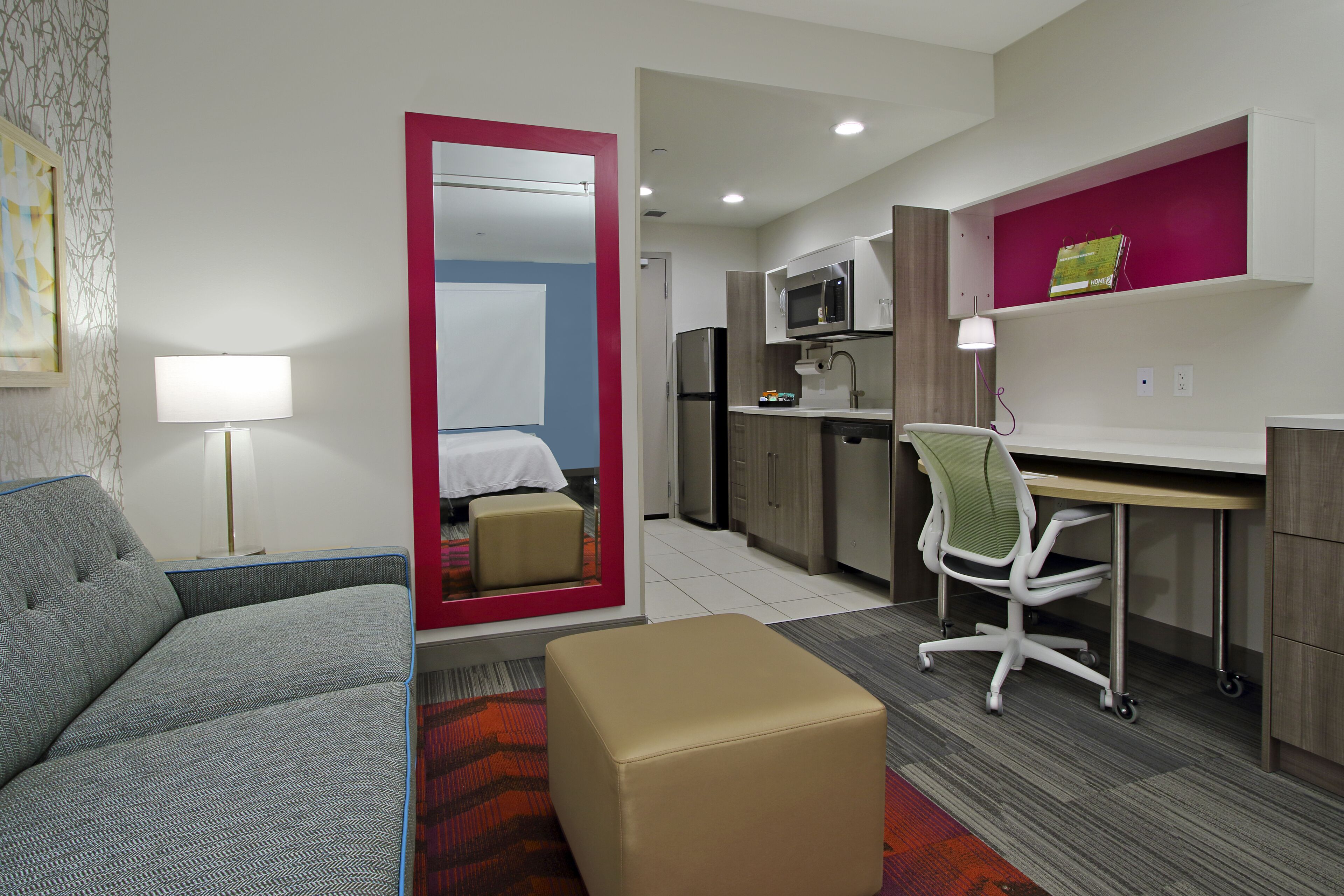 Habitación de hotel compacta con sofá, reposapiés pequeño, escritorio con silla, kitchenette y un espejo que refleja la habitación.