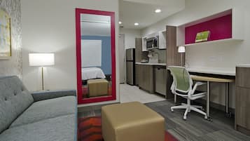 Habitación de hotel compacta con sofá, reposapiés pequeño, escritorio con silla, kitchenette y un espejo que refleja la habitación.